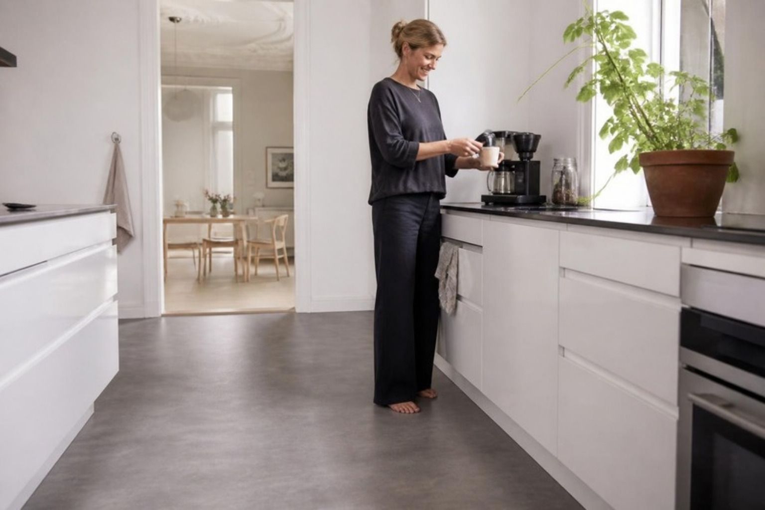 Kvinde laver kaffe i et lyst køkken med hvide skabe og gråt linoleumsgulv